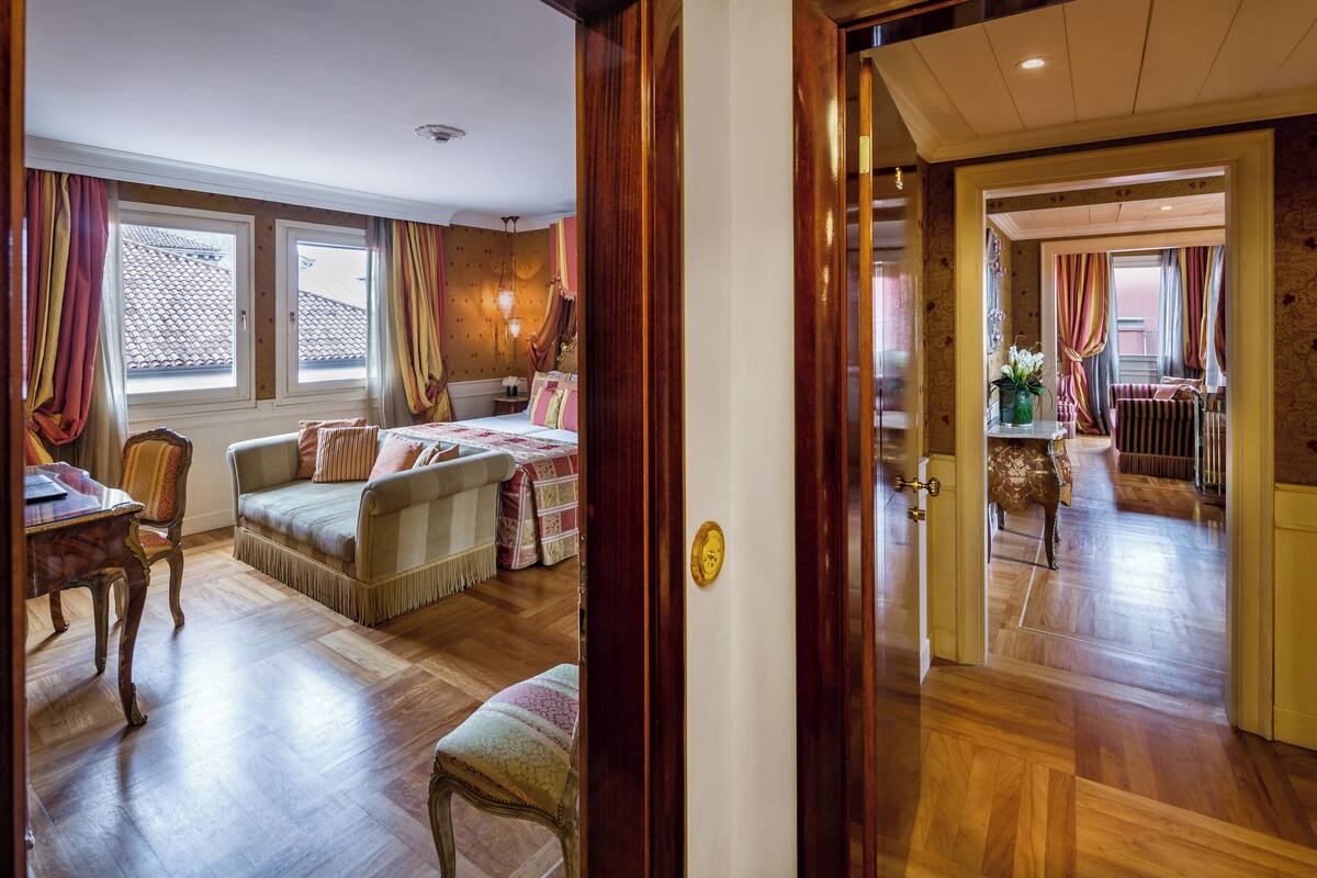 Une chambre d’hôtel avec un plancher en bois, un canapé, une chaise, une table et un lit. Une porte donne accès à une autre pièce.