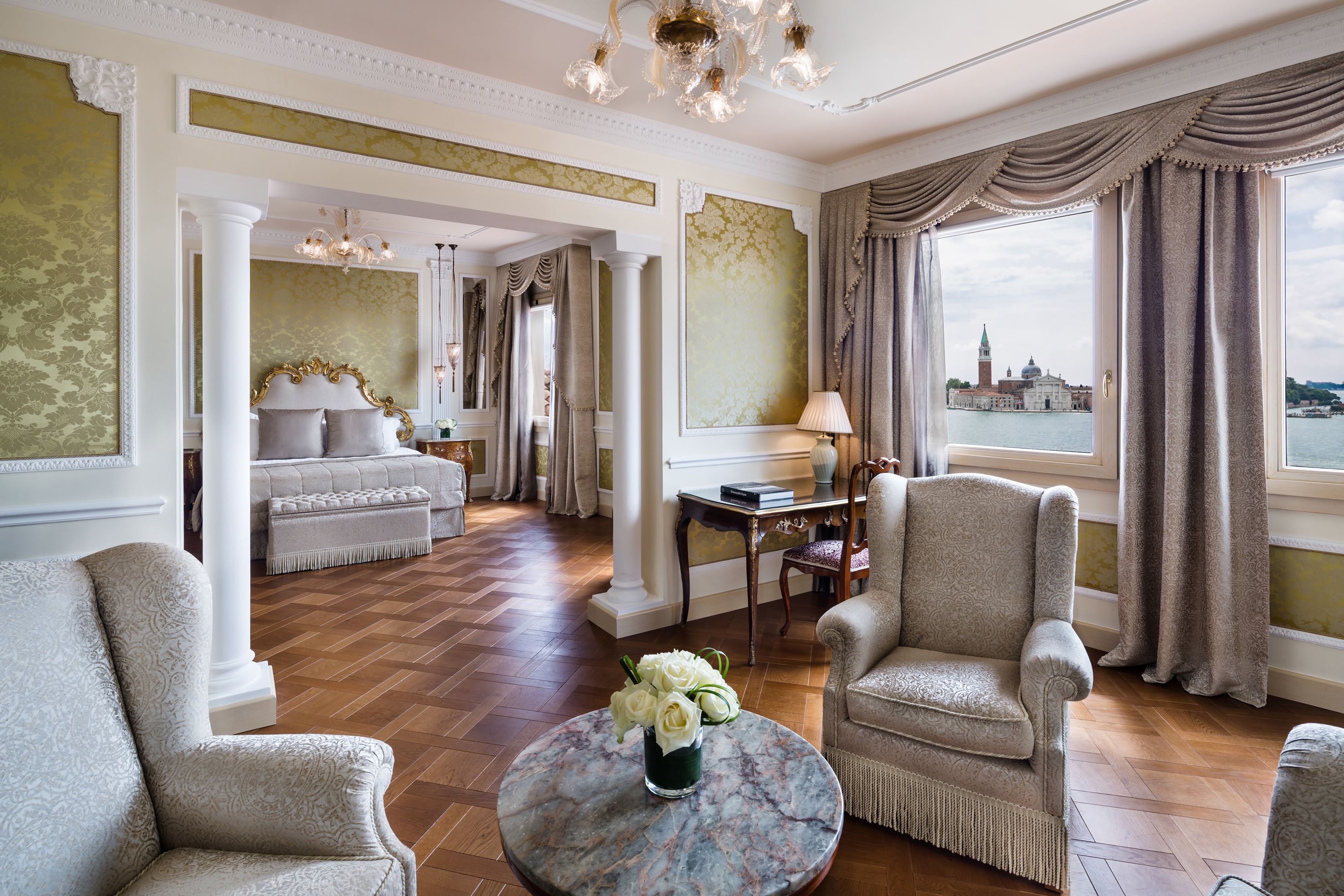 Ett lyxigt hotellrum med en säng, en sittgrupp med bord och stolar, utsikt över stadsbilden genom stora fönster och ett soffbord i marmor med blommor.