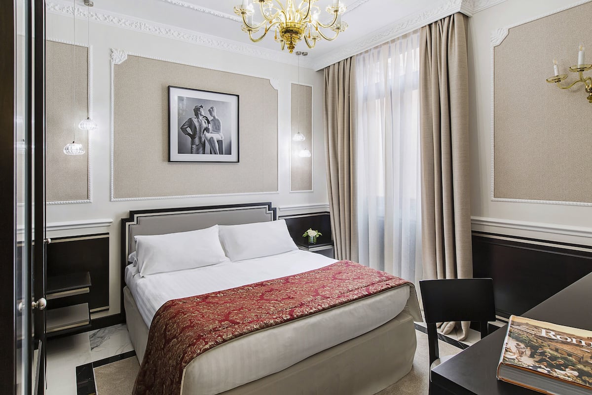 A neatly arranged hotel room with a large bed, a bedside table with a vase, a chair, and a desk with a book.