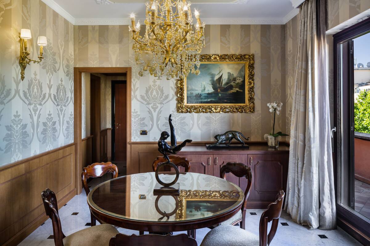 A dining room with a round table, chairs, a chandelier, a painting, and a view through a window.