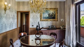 A dining room with a round table, chairs, a chandelier, a painting, and a view through a window.
