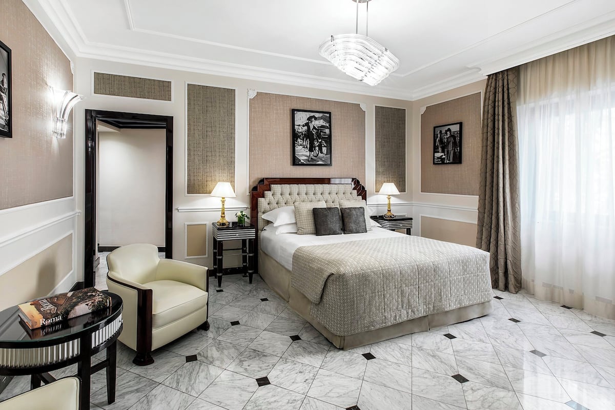 A hotel room with a large bed, a chair, a small table, and two nightstands with lamps.
