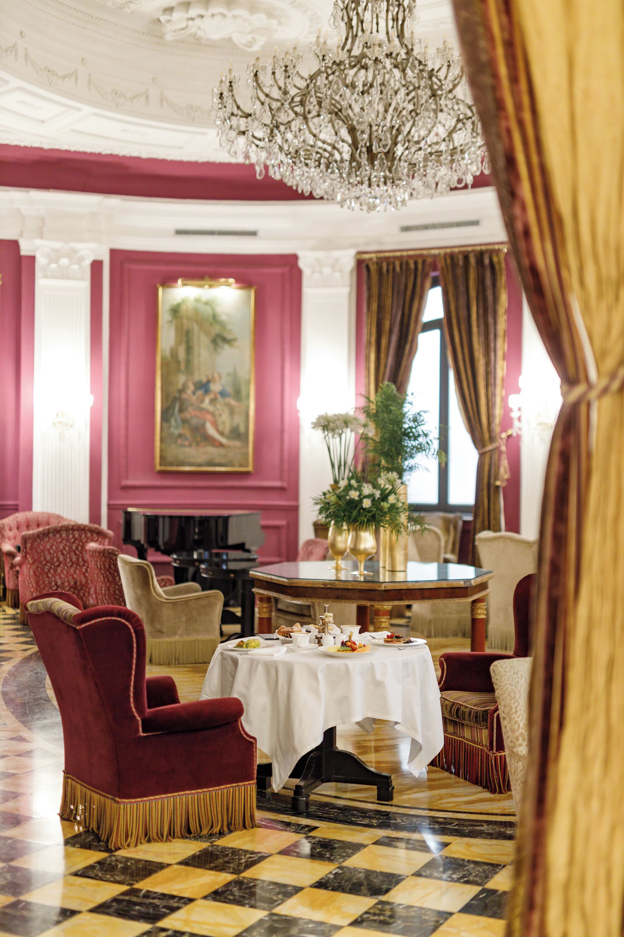 A formal dining room with a chandelier, red upholstered chairs, a round table with a white tablecloth, and a painting on the wall.