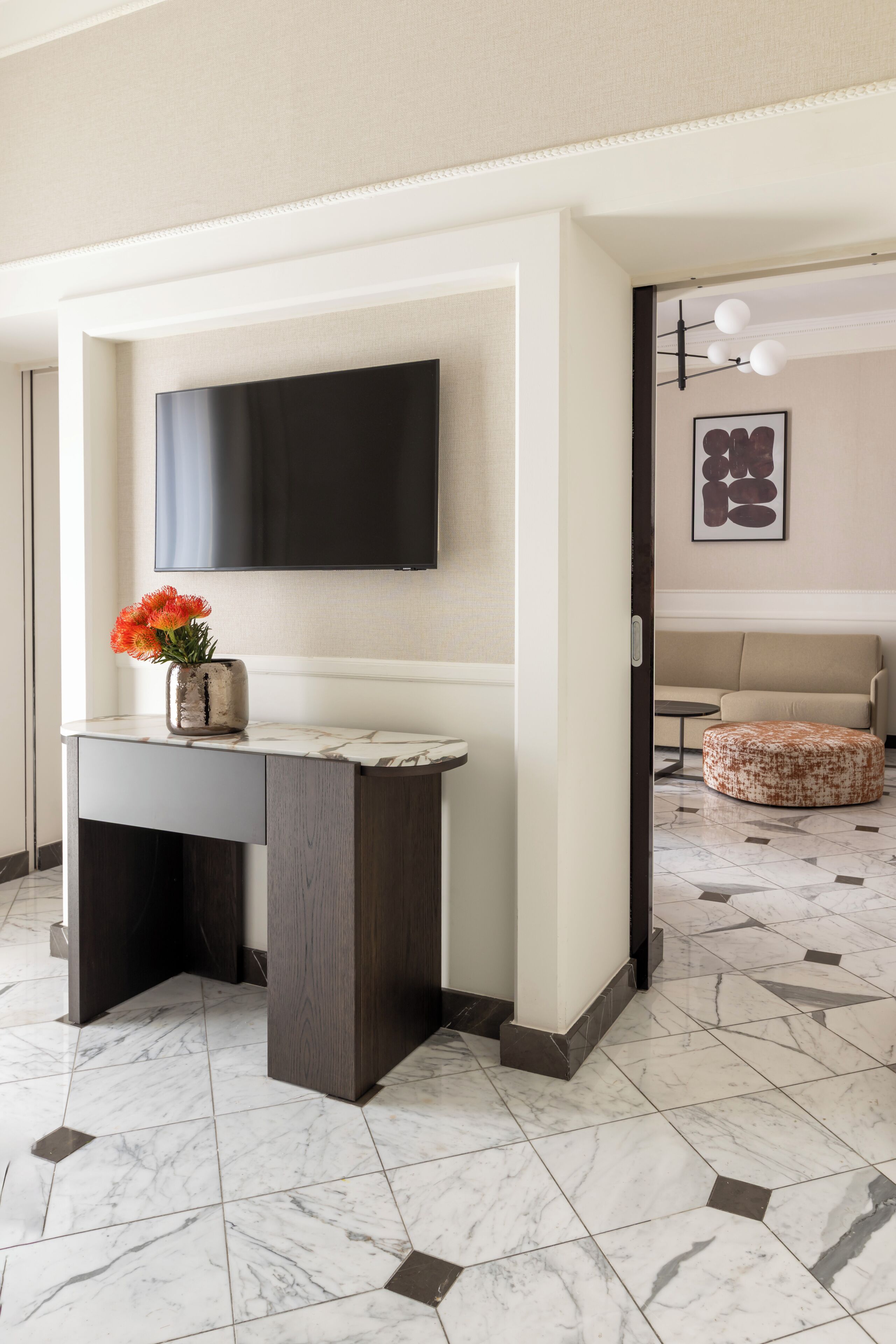 En moderne lobby med et marmorgulv, et mørkt træbord med en blomstervase, et vægmonteret fladskærms-tv og et siddeområde med en mønstret skammel.