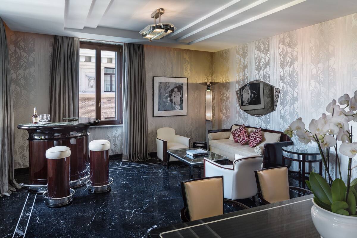 A modern hotel room with a marble floor, a bar area with stools, a sofa, and a coffee table. There is a window with curtains, a framed picture on the wall, and a potted orchid plant.