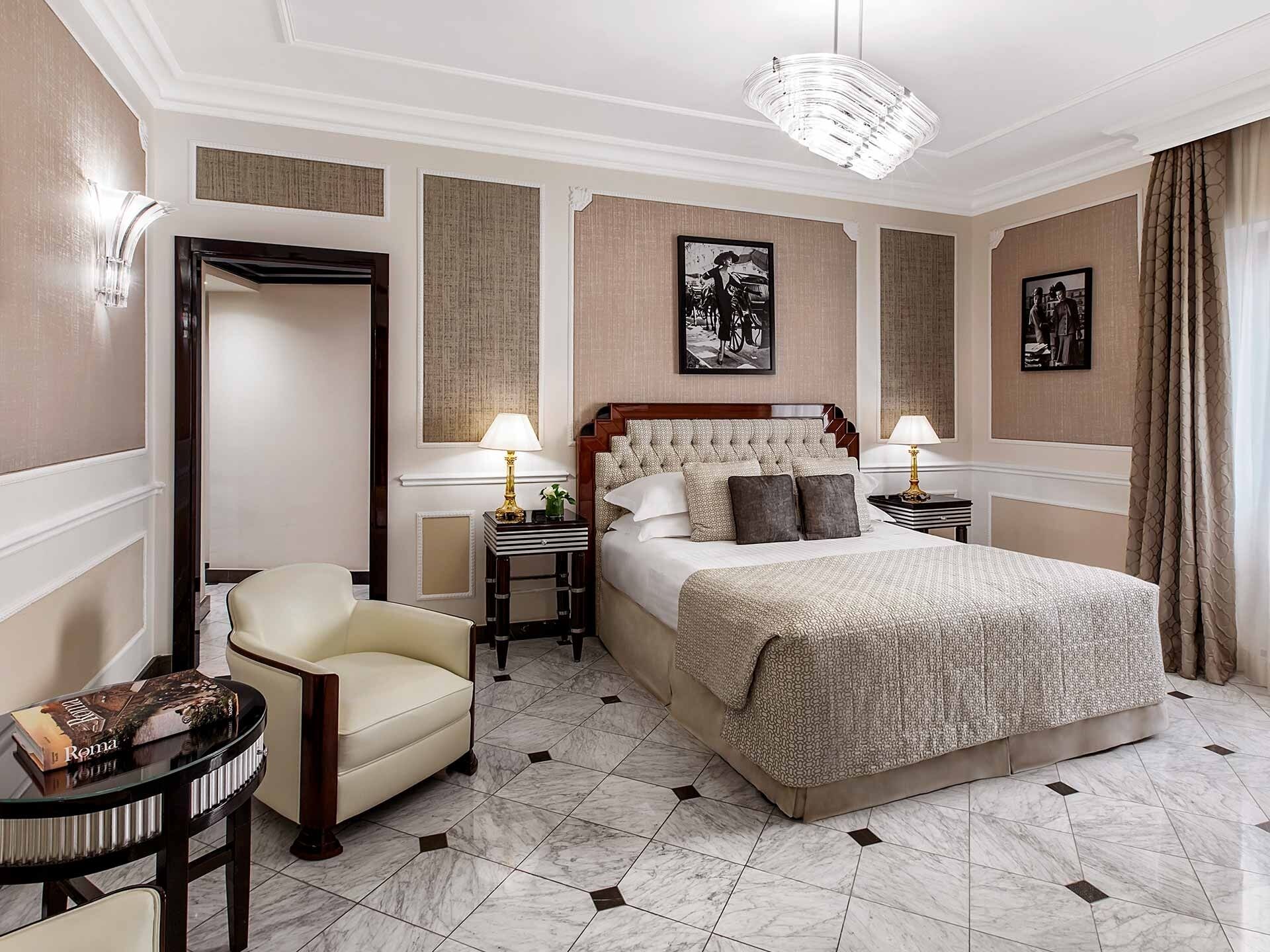 A hotel room with a large bed, two bedside tables, a chair, a mirror, and a chandelier.