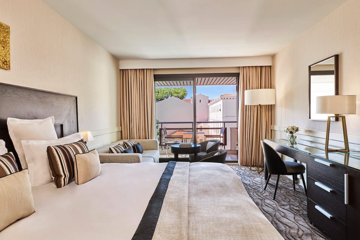 A hotel room with a bed, a desk, a chair, a lamp, and a view of the outside.