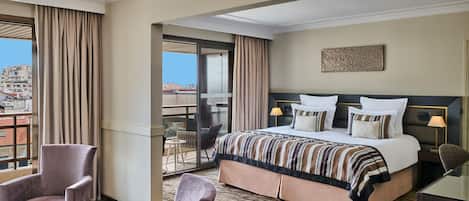 Ein Hotelzimmer mit einem großen Bett, zwei Nachttischen, einem Sessel und Blick auf die Stadt vom Balkon, sowie eine Stadtansicht vom Balkon.