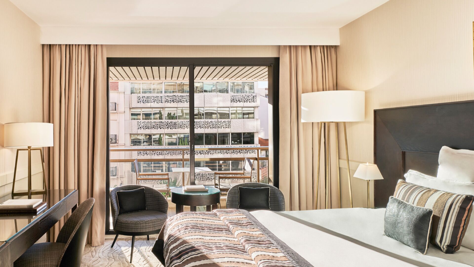Habitación de hotel con cama, dos sillas, una mesita y vista a un edificio con balcones.