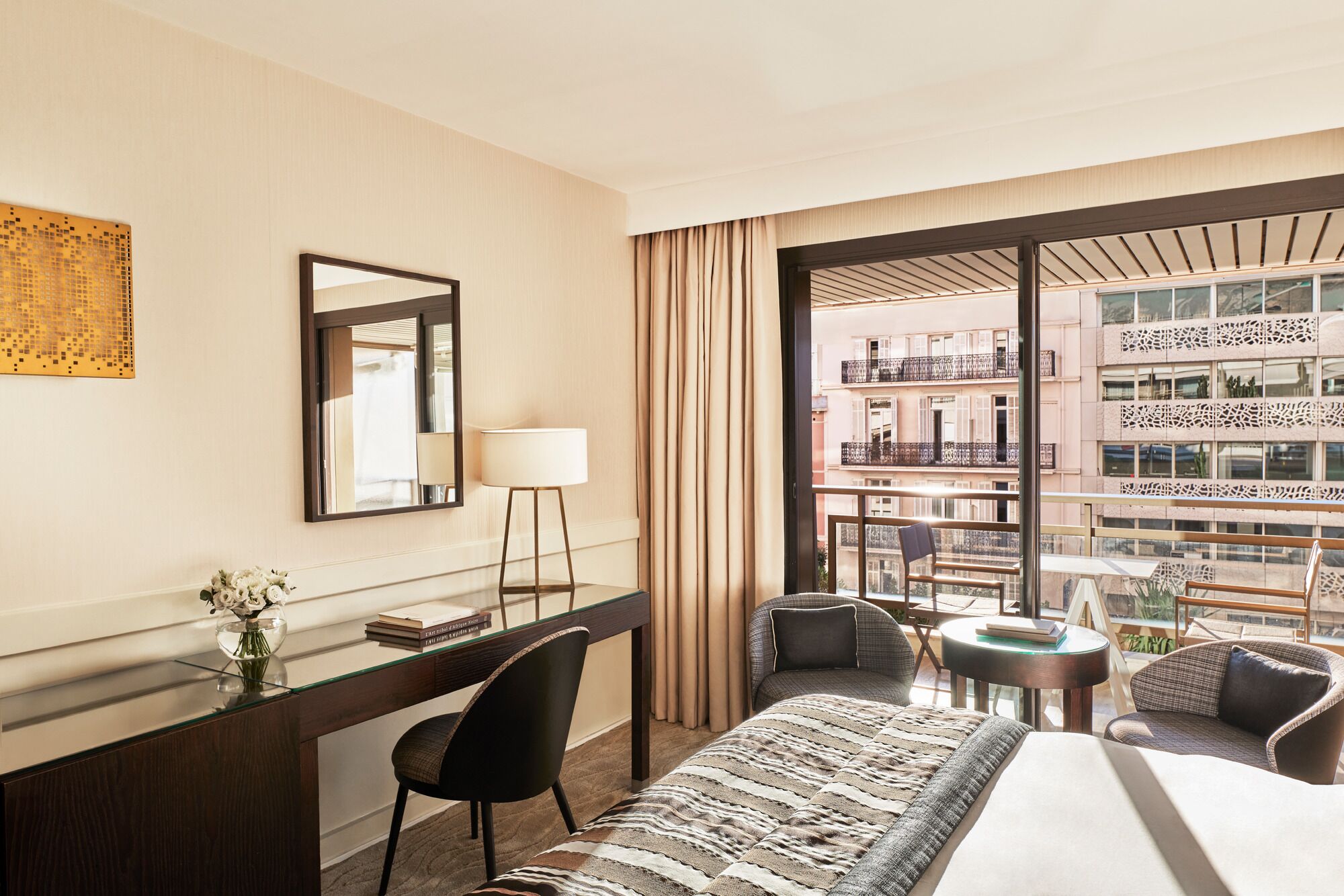 Habitación de hotel con una cama, un escritorio con silla, una pequeña mesa con silla, un ventanal con cortinas y vista a un edificio con balcones.