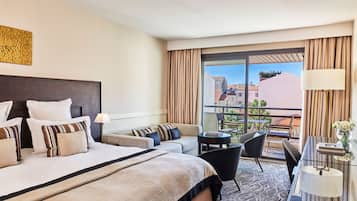 Une chambre d’hôtel avec un grand lit, un canapé, un bureau avec une lampe et un balcon donnant sur des immeubles.