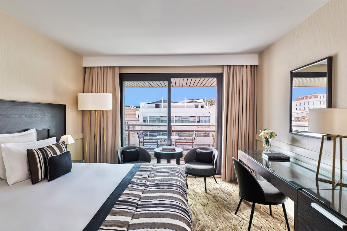 A hotel room with a bed, a desk, a chair, a small table, and a large window with curtains.