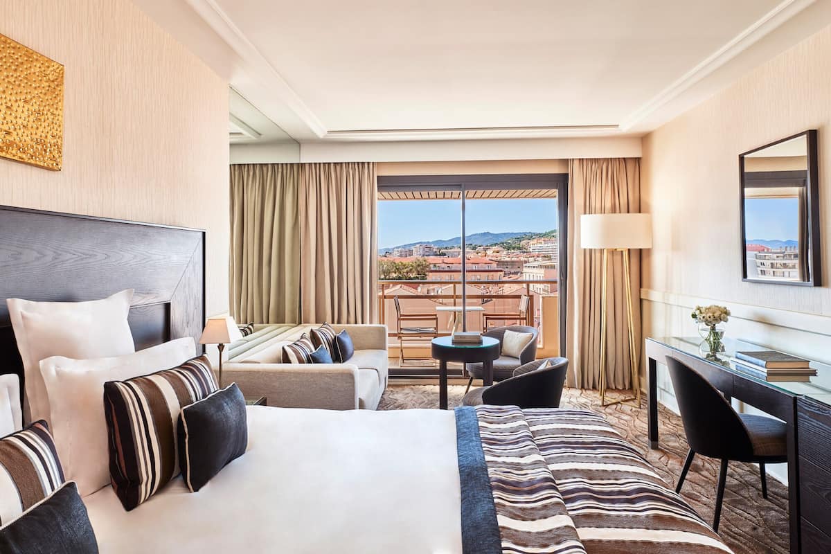 A hotel room with a large bed, a desk, a chair, a sofa, and a view of the city.