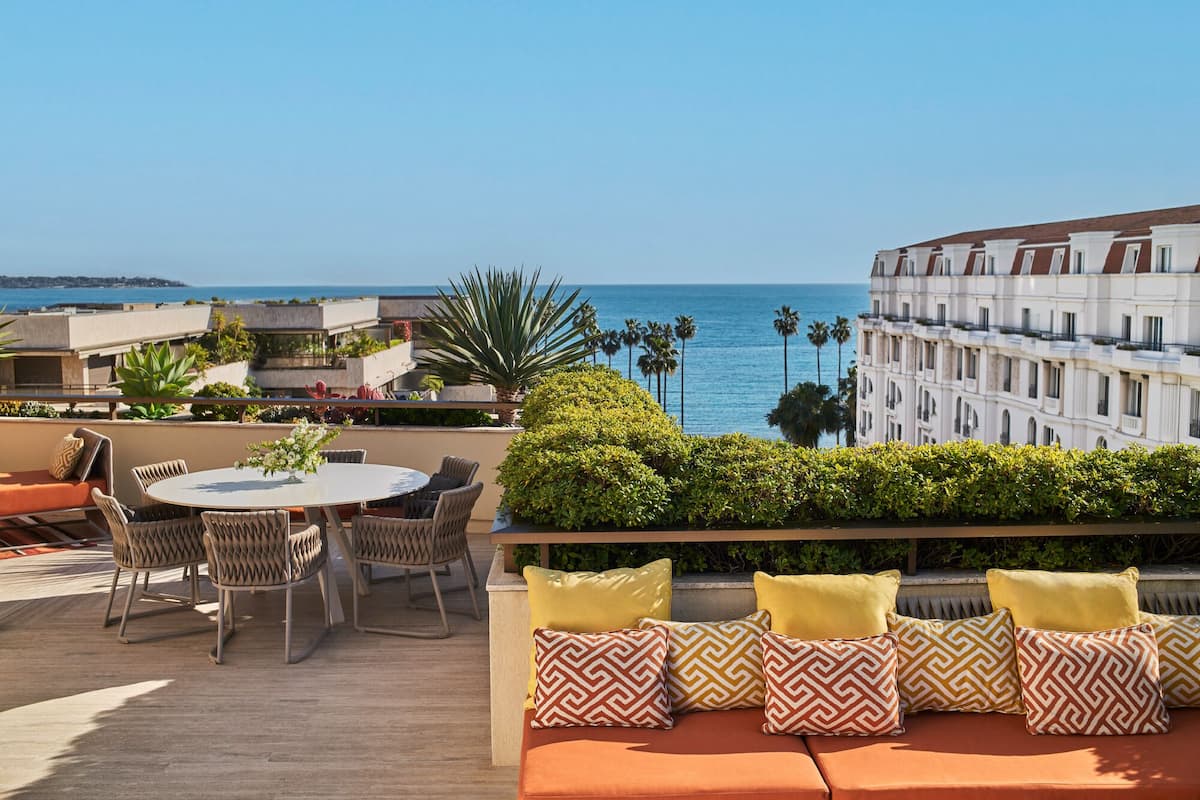 A terrace with a round table and chairs, a sofa with patterned cushions, and a view of the ocean and palm trees.