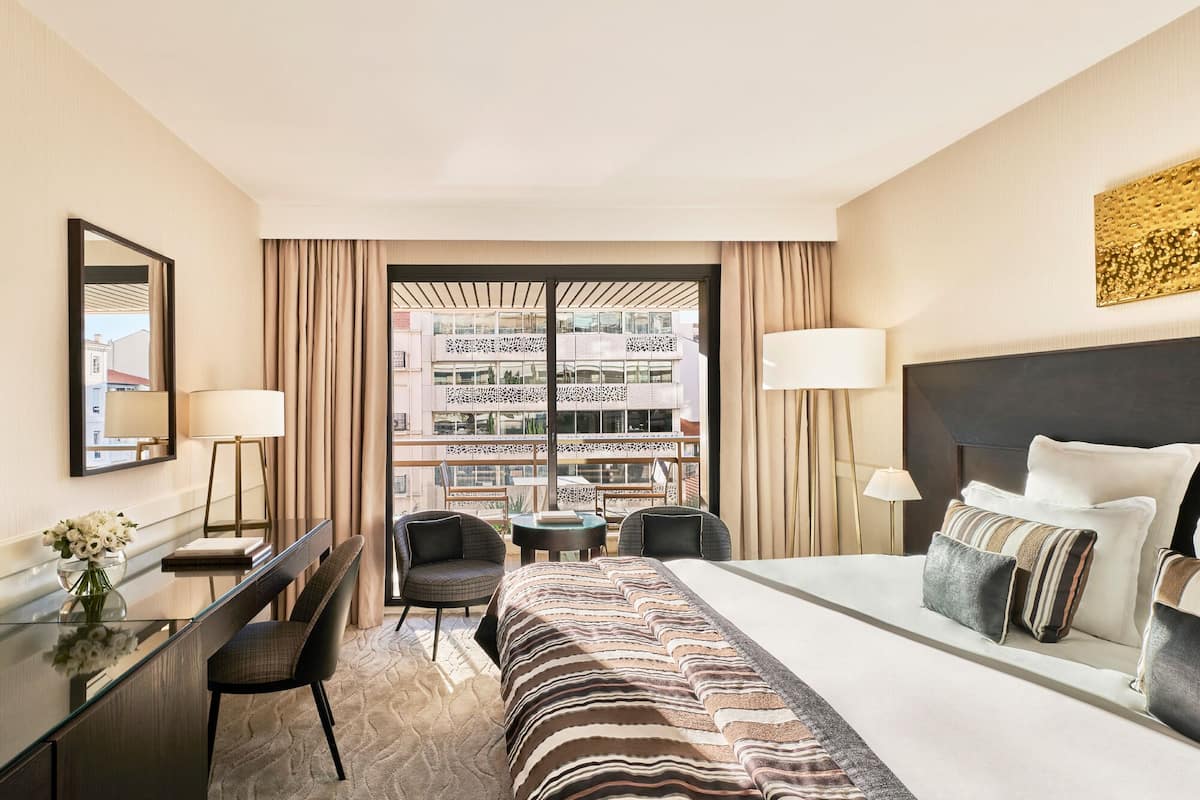 A hotel room with a large bed, a desk with a chair, a small table, and a view of a building through a window with curtains.