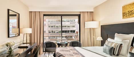 A hotel room with a large bed, a desk with a chair, a small table, and a view of a building through a window with curtains.