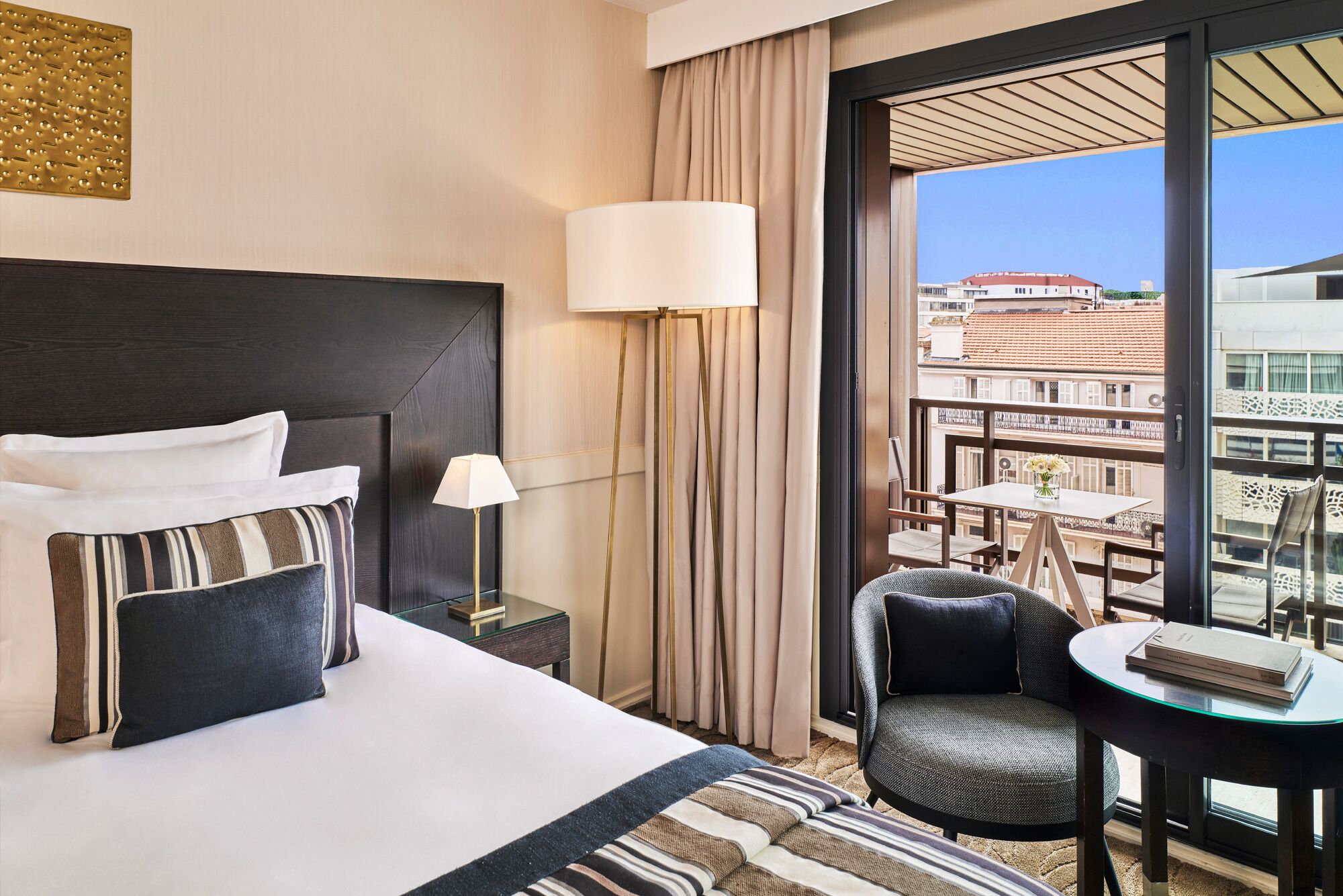A hotel room with a bed, bedside table, lamp, and a view of the city through large windows.