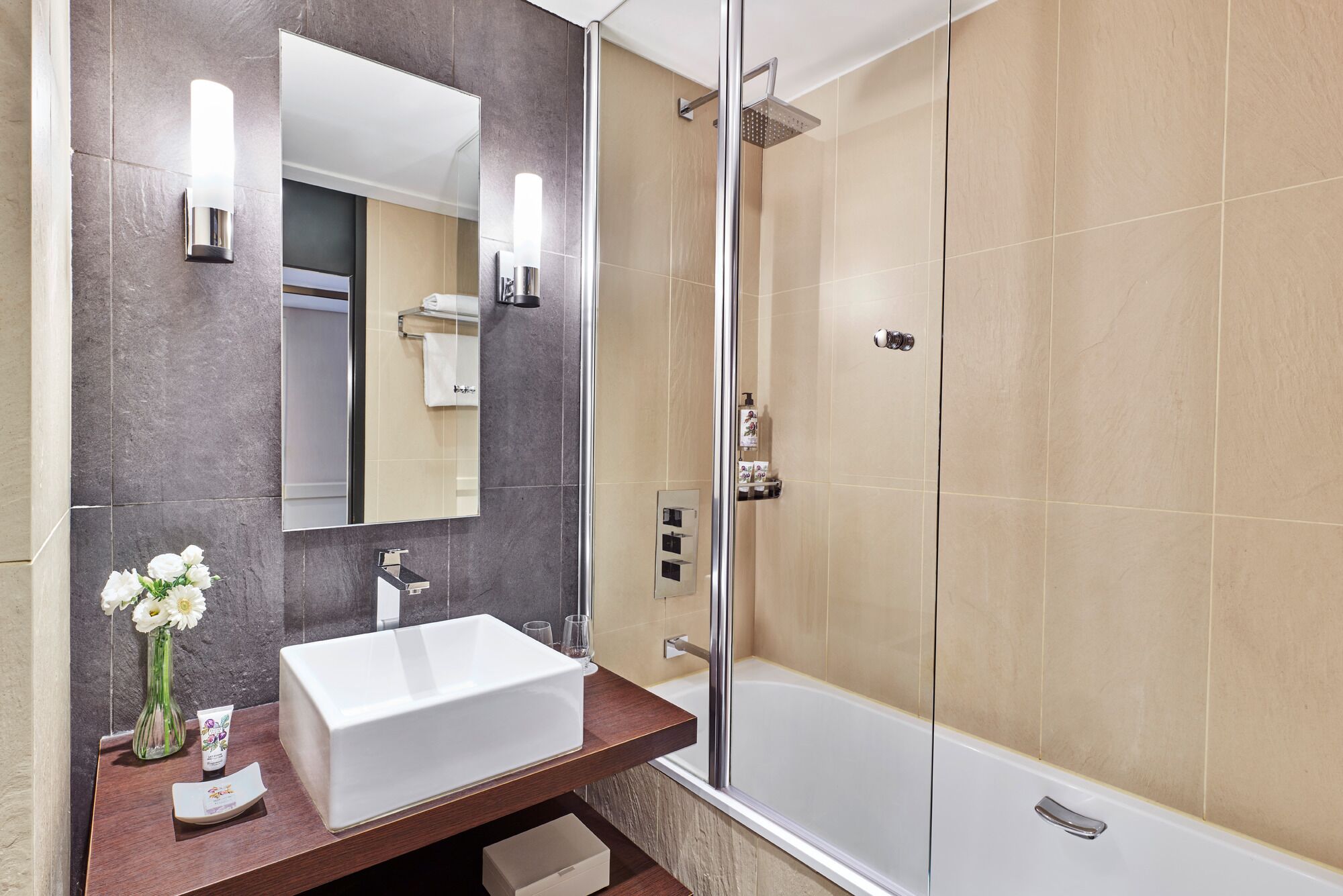 A modern bathroom with a large mirror, a white sink, a shower, and a vase of flowers.