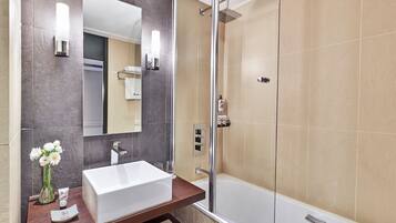 A modern bathroom with a large mirror, a white sink, a shower, and a vase of flowers.