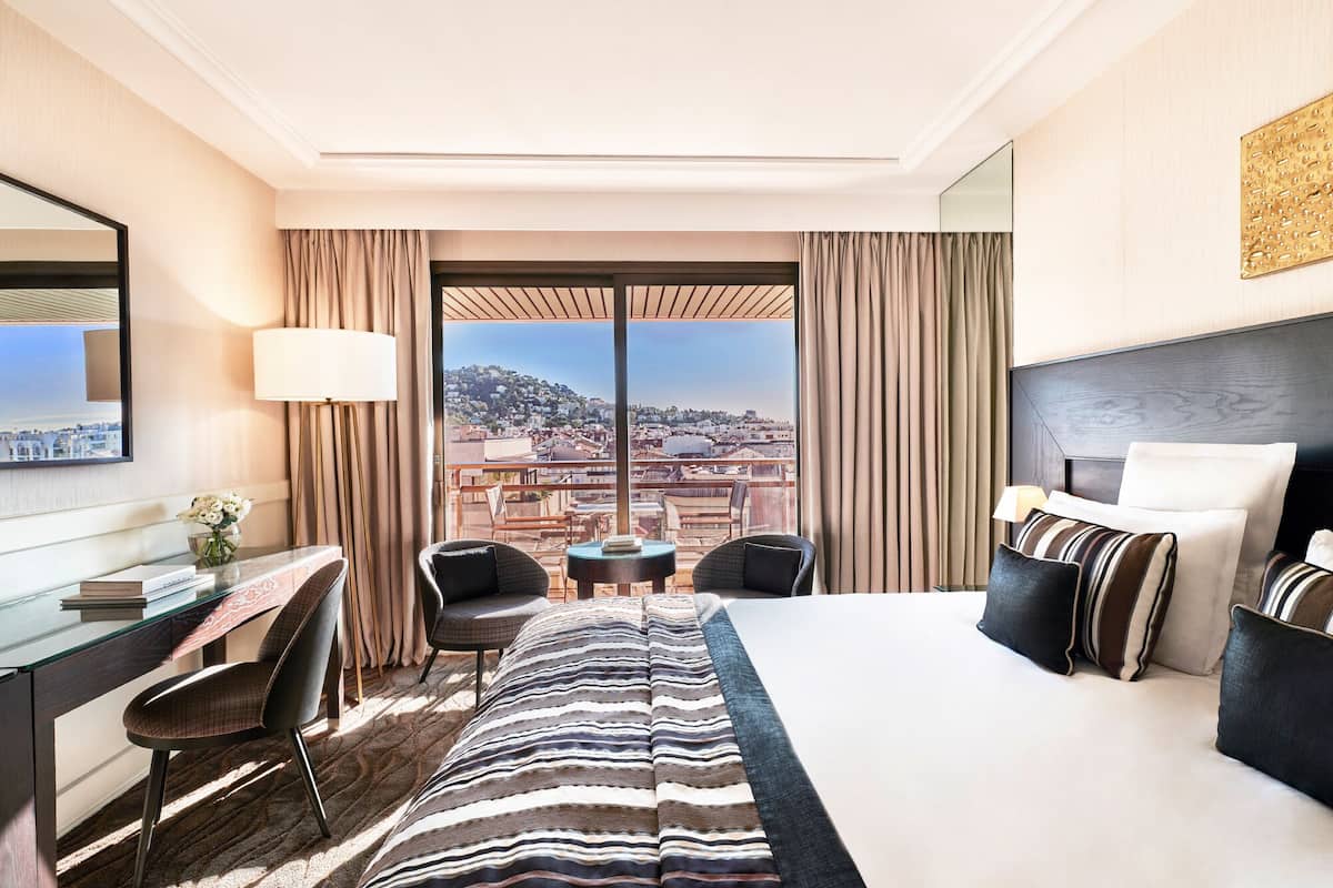 A hotel room with a large bed, a desk with a chair, a small table with two chairs, and a view of a cityscape through large windows.