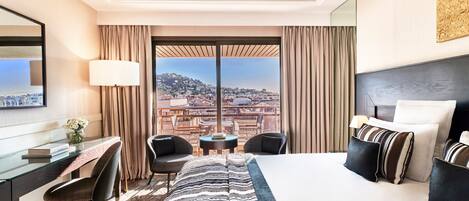 Habitación de hotel con una cama grande, un escritorio con una silla, una pequeña mesa con dos sillas y vistas a un paisaje urbano a través de amplios ventanales.