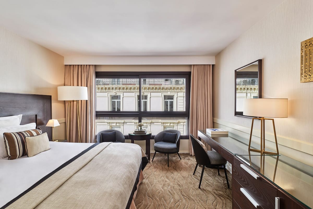 A hotel room with a bed, desk, chair, and a view of a building through the window.