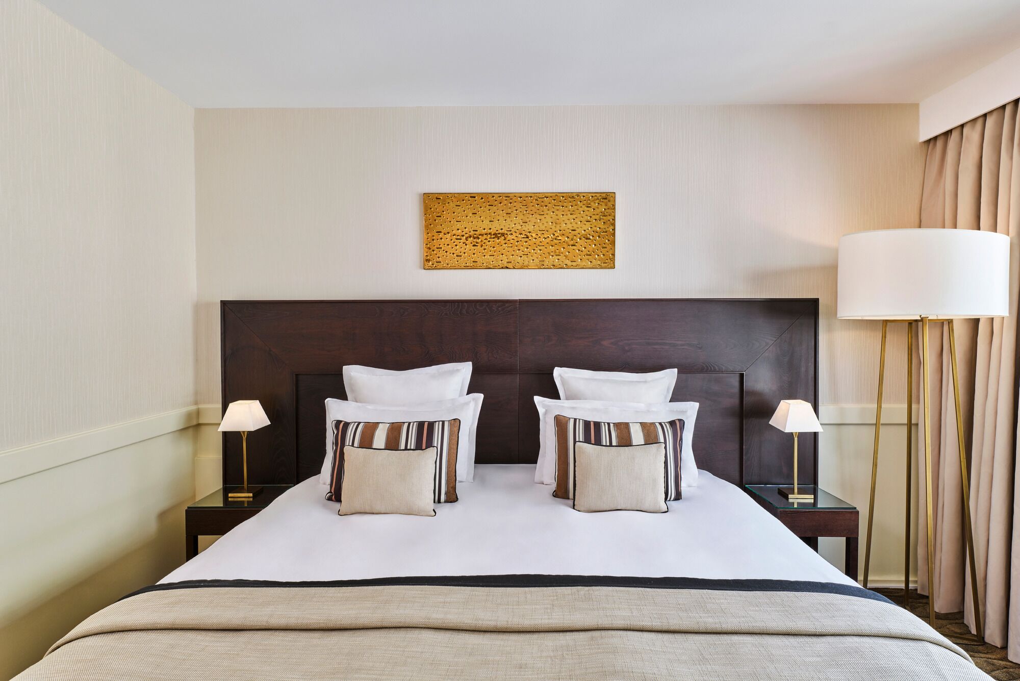 A hotel room with a large bed, two bedside tables, a headboard, a wall-mounted artwork, and a window with curtains.