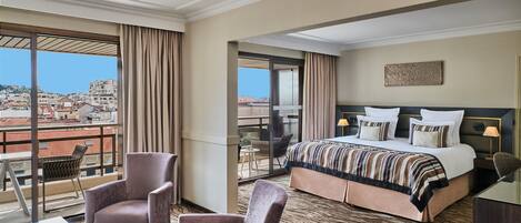 A hotel room with a large bed, two armchairs, a small table, and a view of the cityscape.