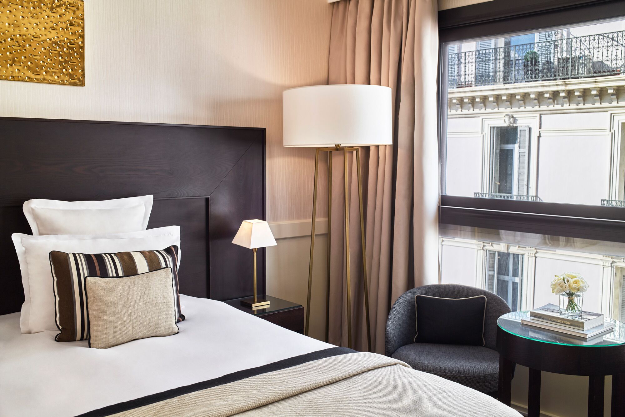 A hotel room with a large bed, a bedside table, a lamp, a chair, and a view of the city through a window.