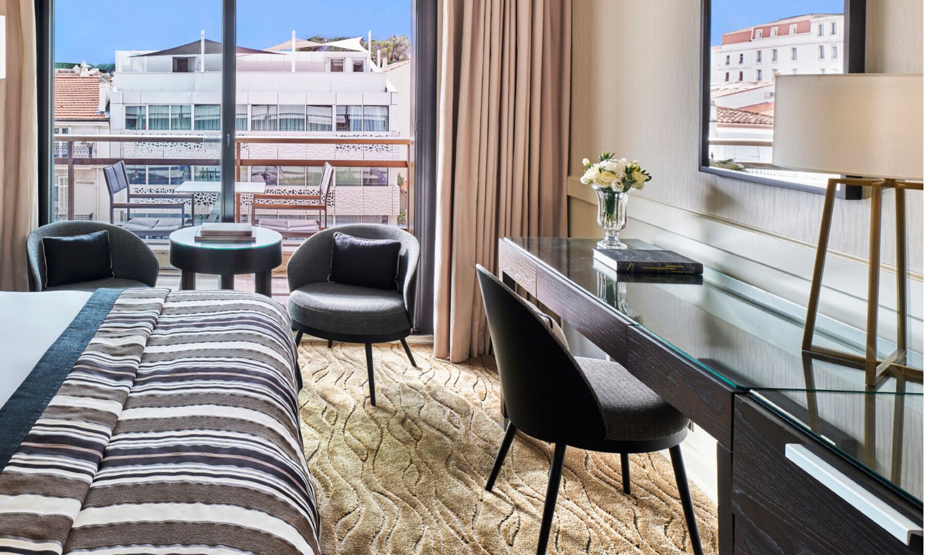 Une chambre d’hôtel comprenant un lit, deux fauteuils, une petite table ronde, un bureau avec une chaise, un vase de fleurs et une vue sur les bâtiments à travers de grandes fenêtres.