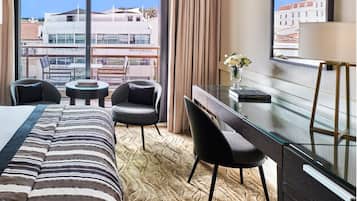 A hotel room with a bed, two armchairs, a small round table, a desk with a chair, a vase with flowers, and a view of buildings through large windows.
