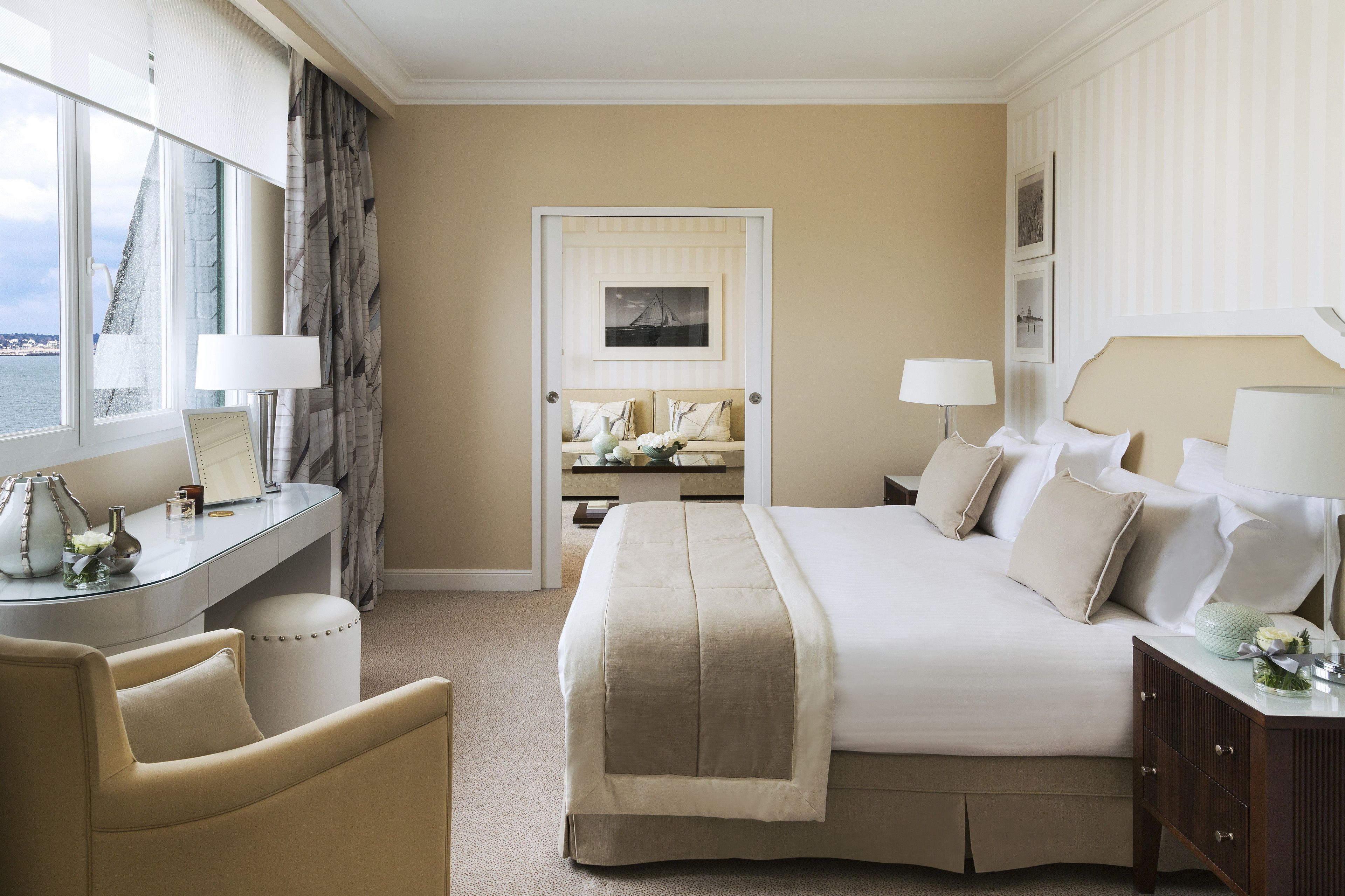 A hotel room with a large bed, a desk with a lamp, a chair, and a view of the sea through the window.