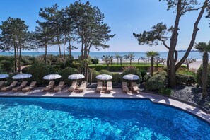 Une piscine entourée d’arbres, avec des chaises longues et des parasols, offrant une vue sur la mer.