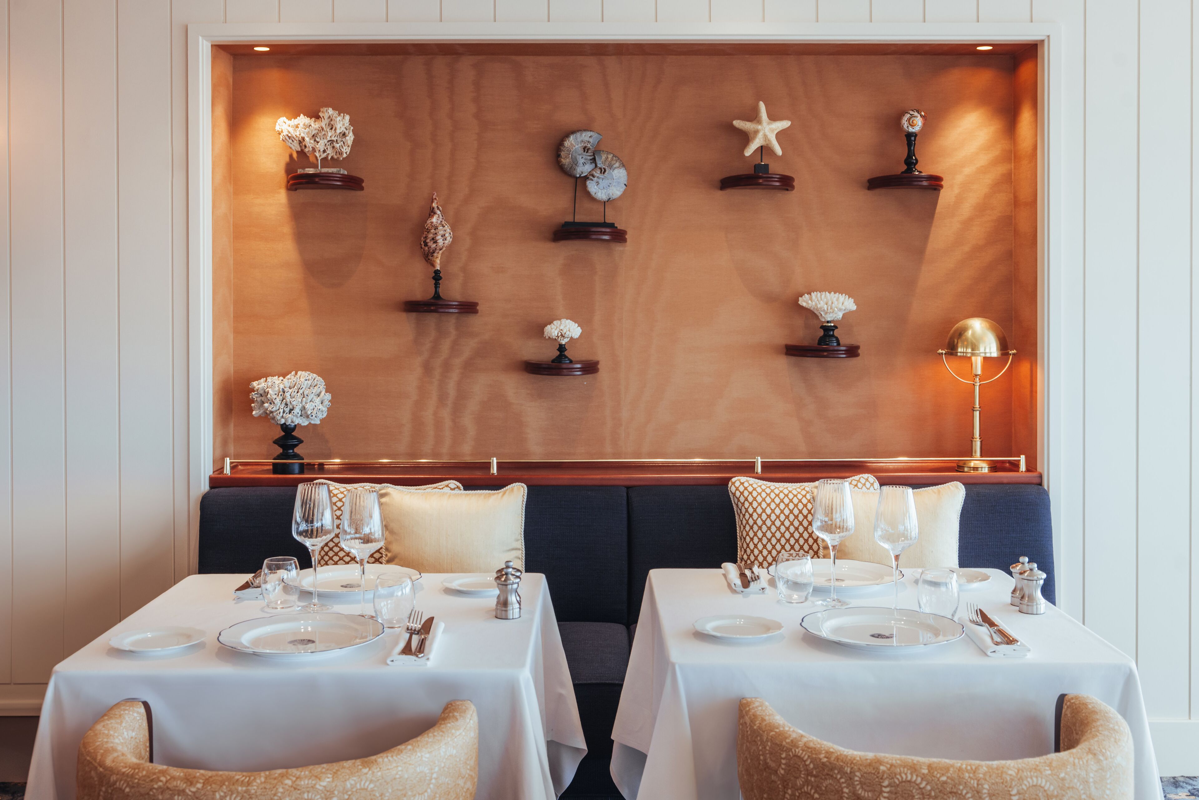 A dining area with two tables set for two, each with white tablecloths, silverware, and glassware. The tables are flanked by cushioned chairs. A wooden wall with decorative items and a lamp is behind the tables.