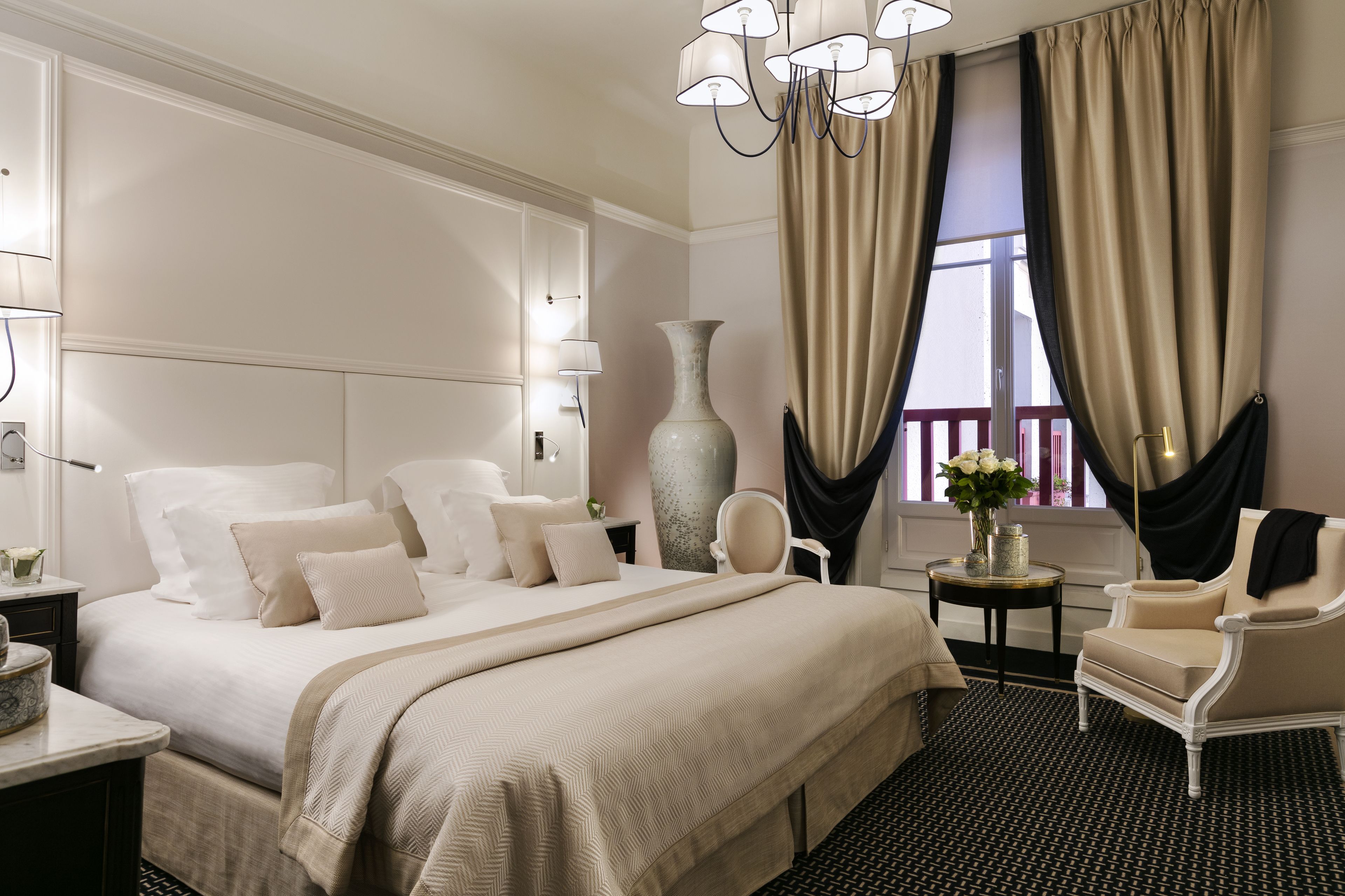 A hotel room with a large bed, a sitting area with a chair, a vase with flowers, and a large window with curtains.