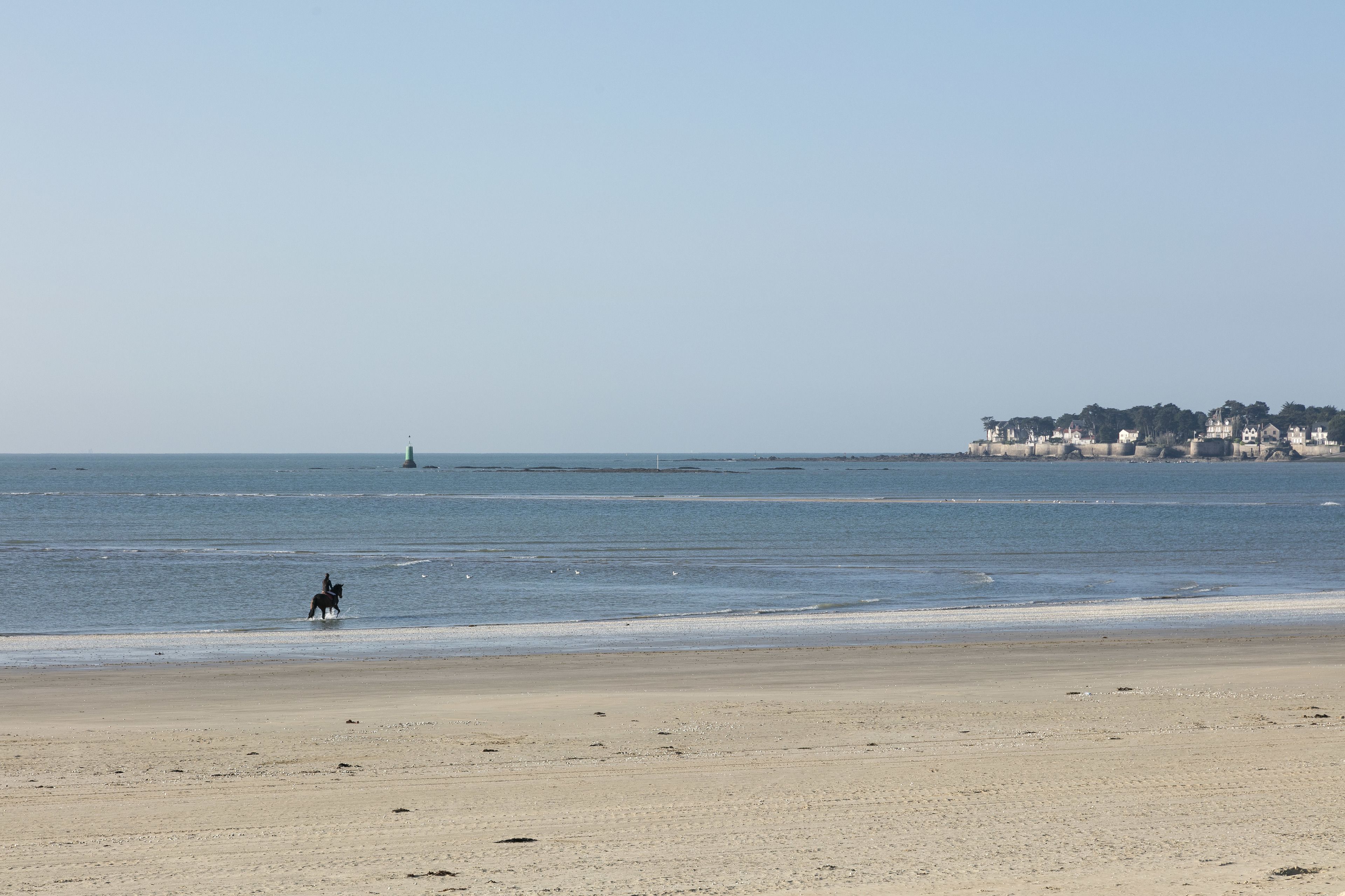 A beach with a person riding a horse, a lighthouse in the distance, and a building on the shore.