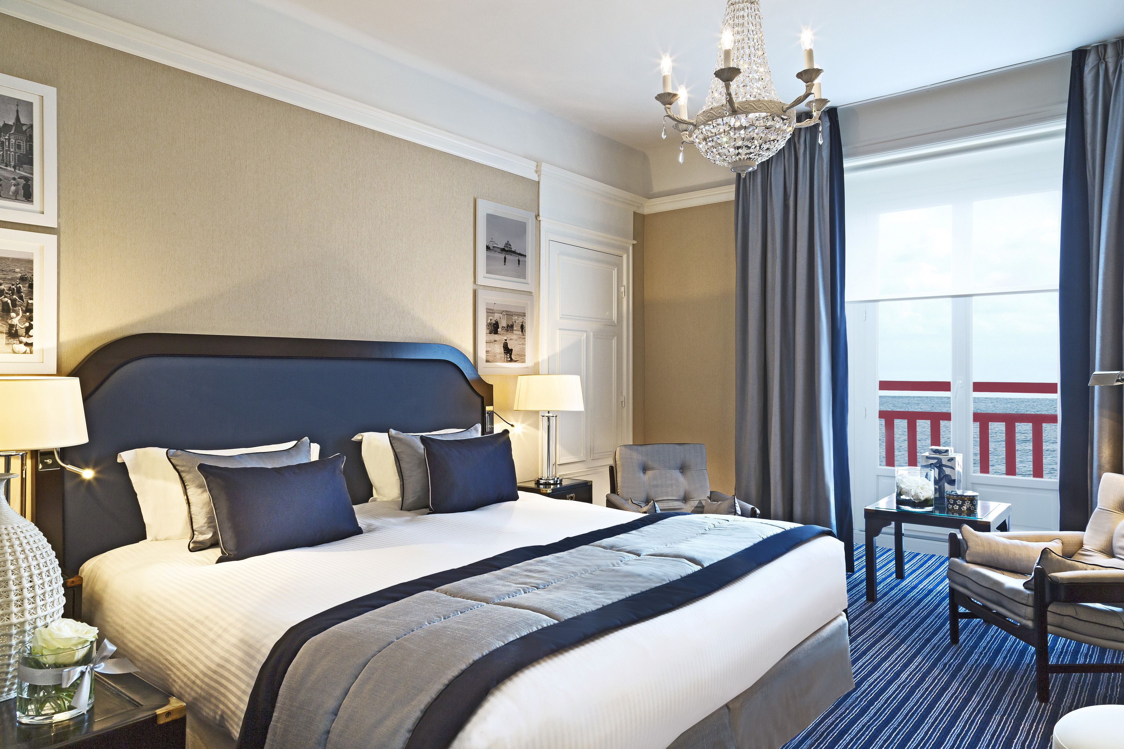 Habitación de hotel con una cama grande, un candelabro, vista a un balcón y un escritorio con lámpara.