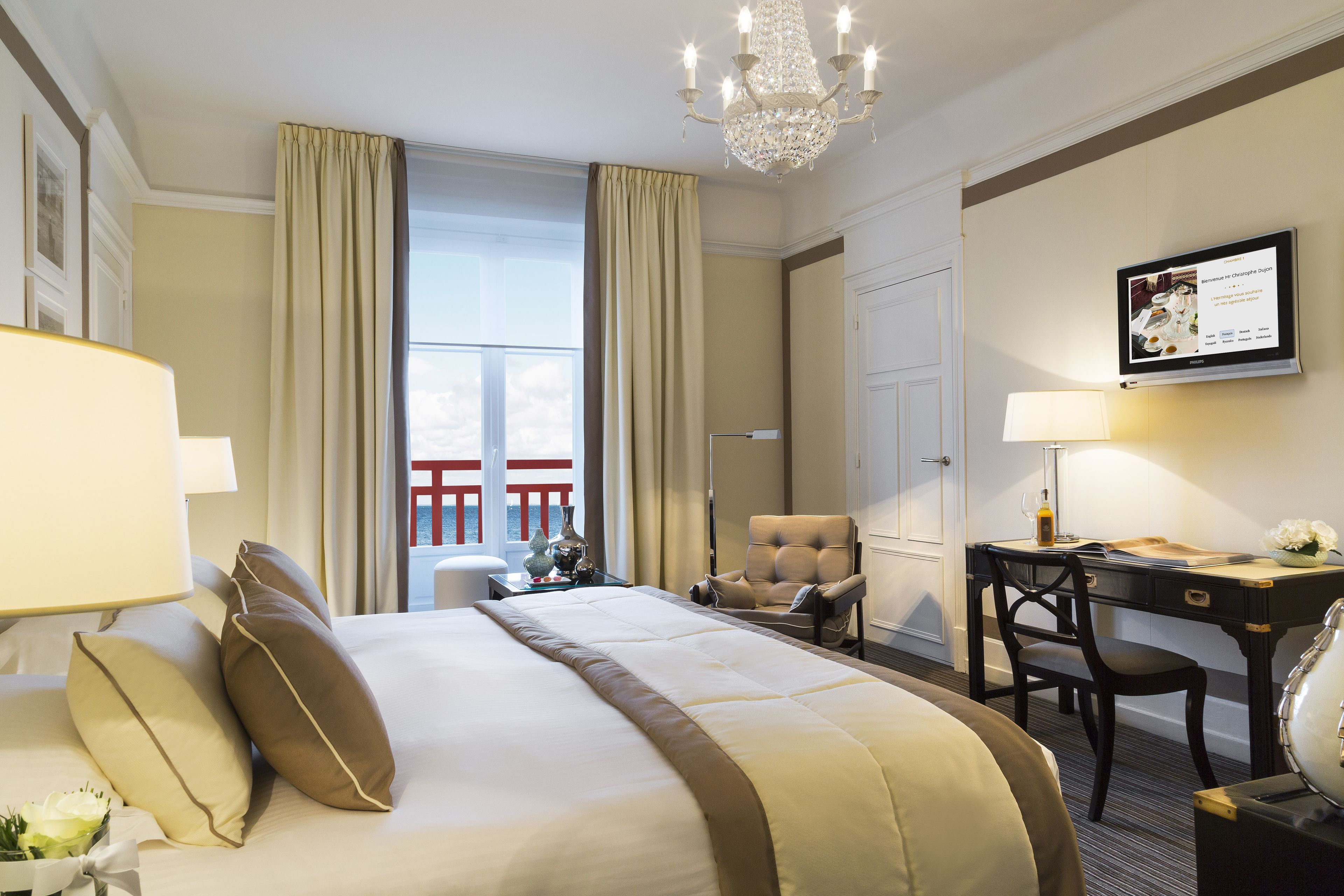A hotel room with a large bed, a desk with a chair, a television, and a chandelier.