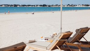 Un espai relaxant vora la platja amb cadires de fusta i una sombrereta blanca, amb vista a un mar blau i una costa llunyana.