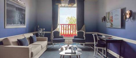 A room with a sofa, two chairs, a table, a chandelier, a window with curtains, and a wall-mounted light fixture.