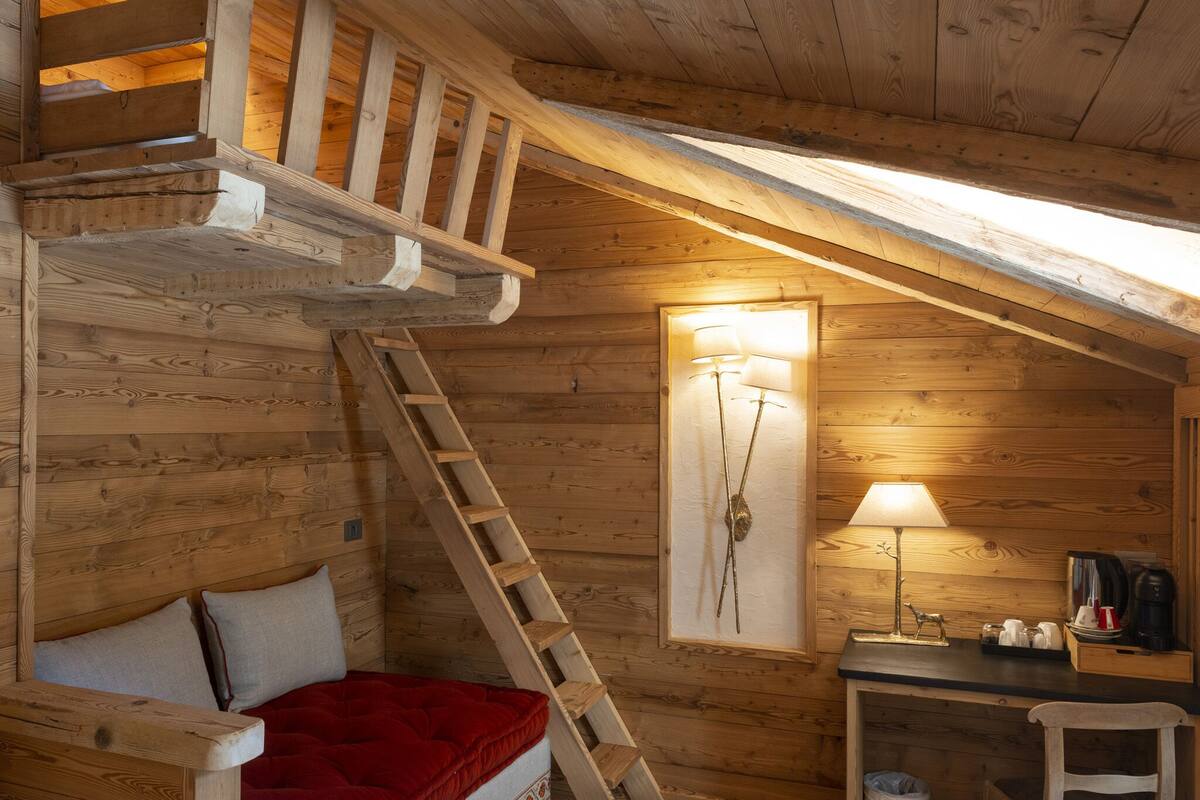 A cozy cabin room with a loft bed, a wooden ladder, a desk with a lamp, and a framed picture on the wall.