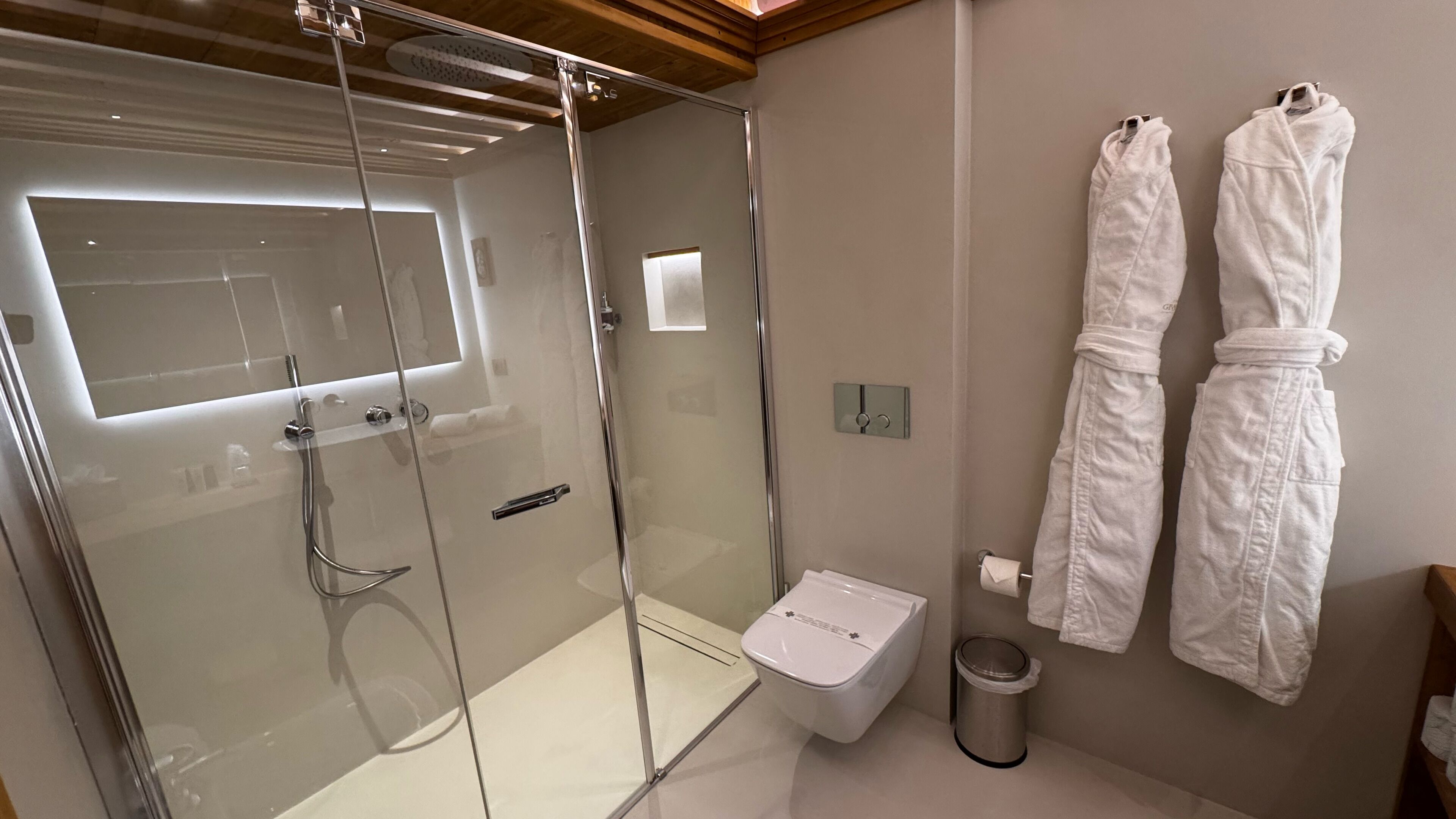 A modern bathroom with a glass-enclosed shower, a toilet, and two white robes hanging on the wall.