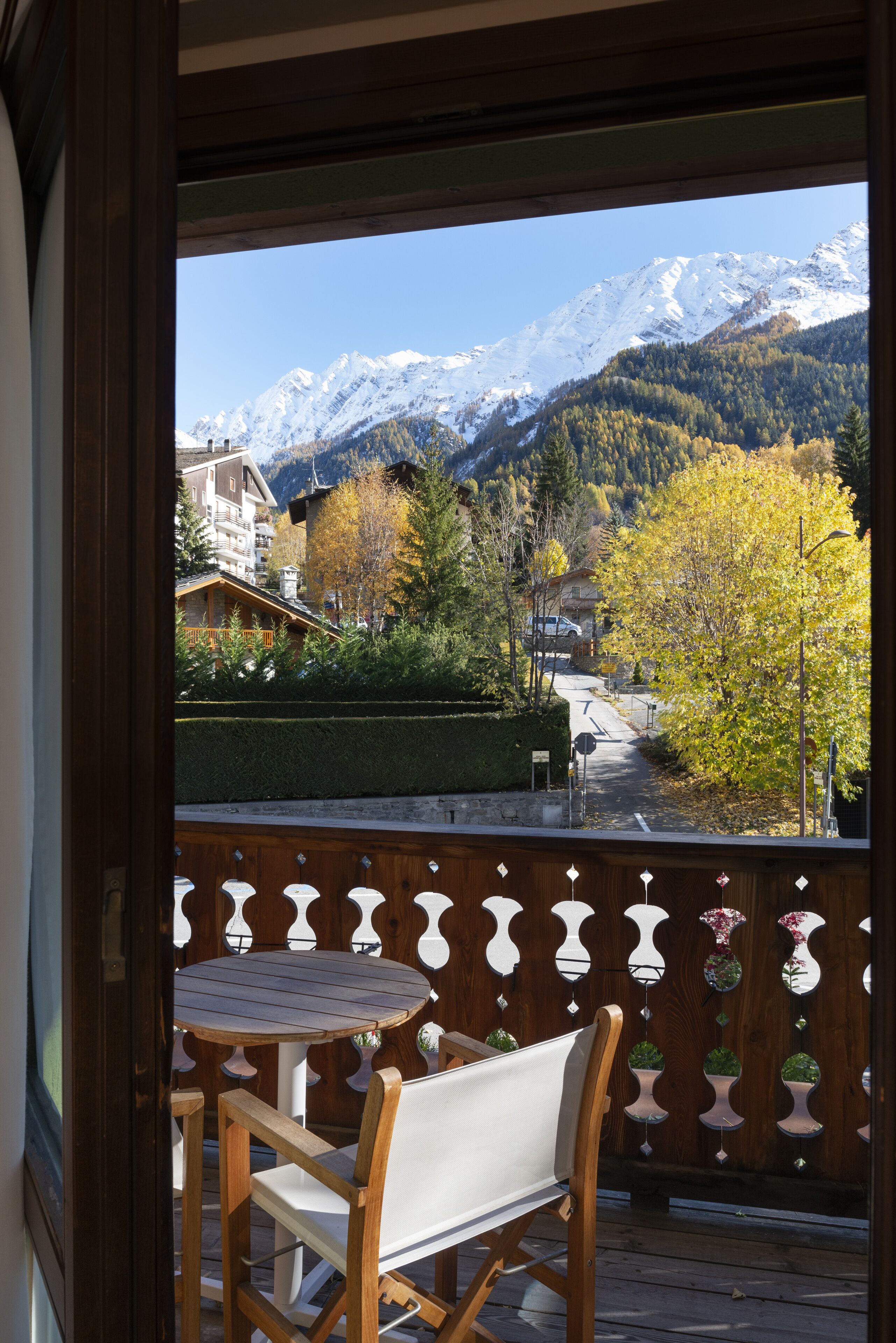 Eine Holzterrasse mit einem runden Tisch und zwei Stühlen, mit Blick auf eine Berglandschaft mit schneebedeckten Gipfeln und herbstlich gefärbten Bäumen.