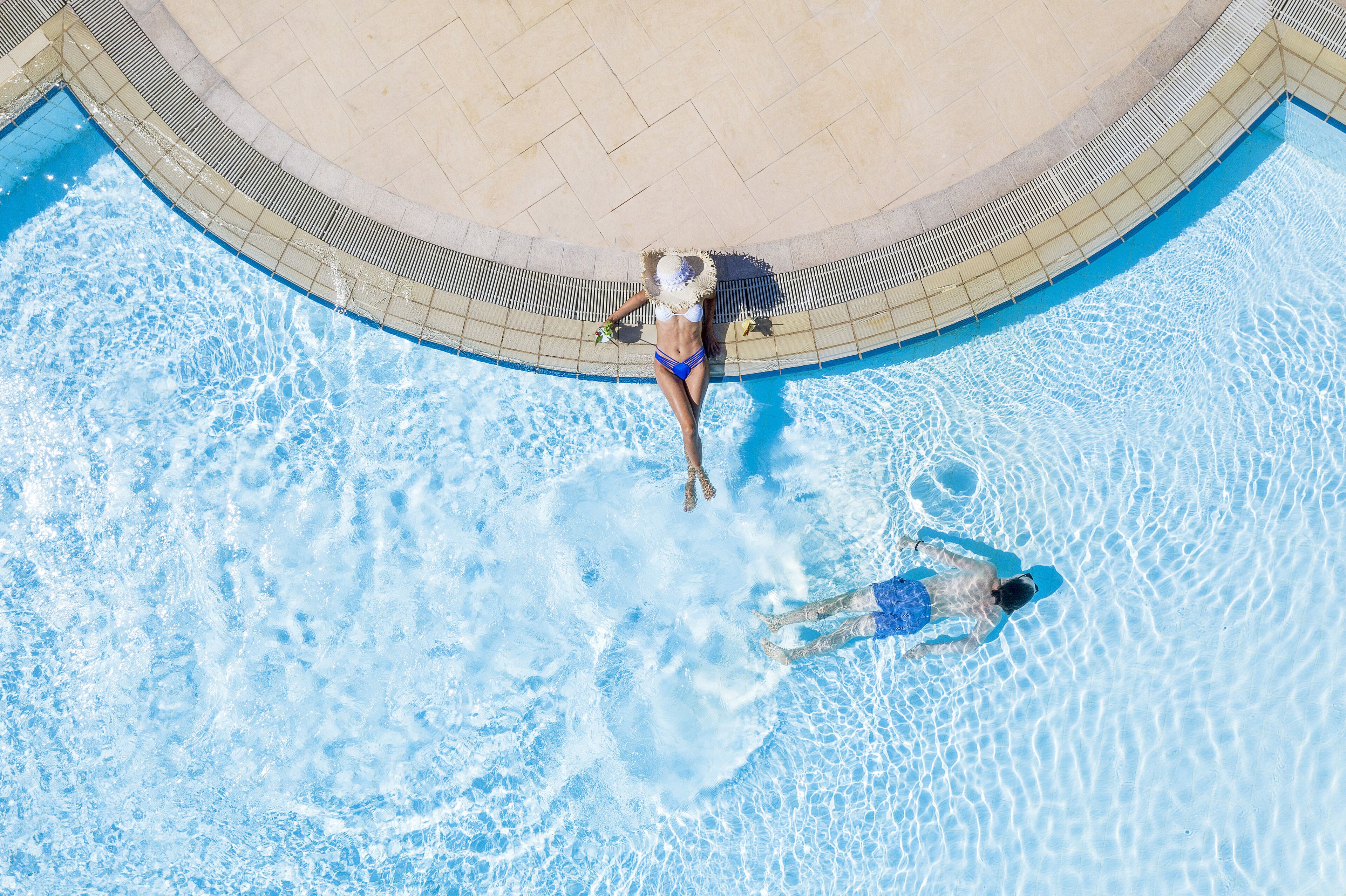 An aerial view of a swimming pool with two people in the water, surrounded by a tiled deck.
