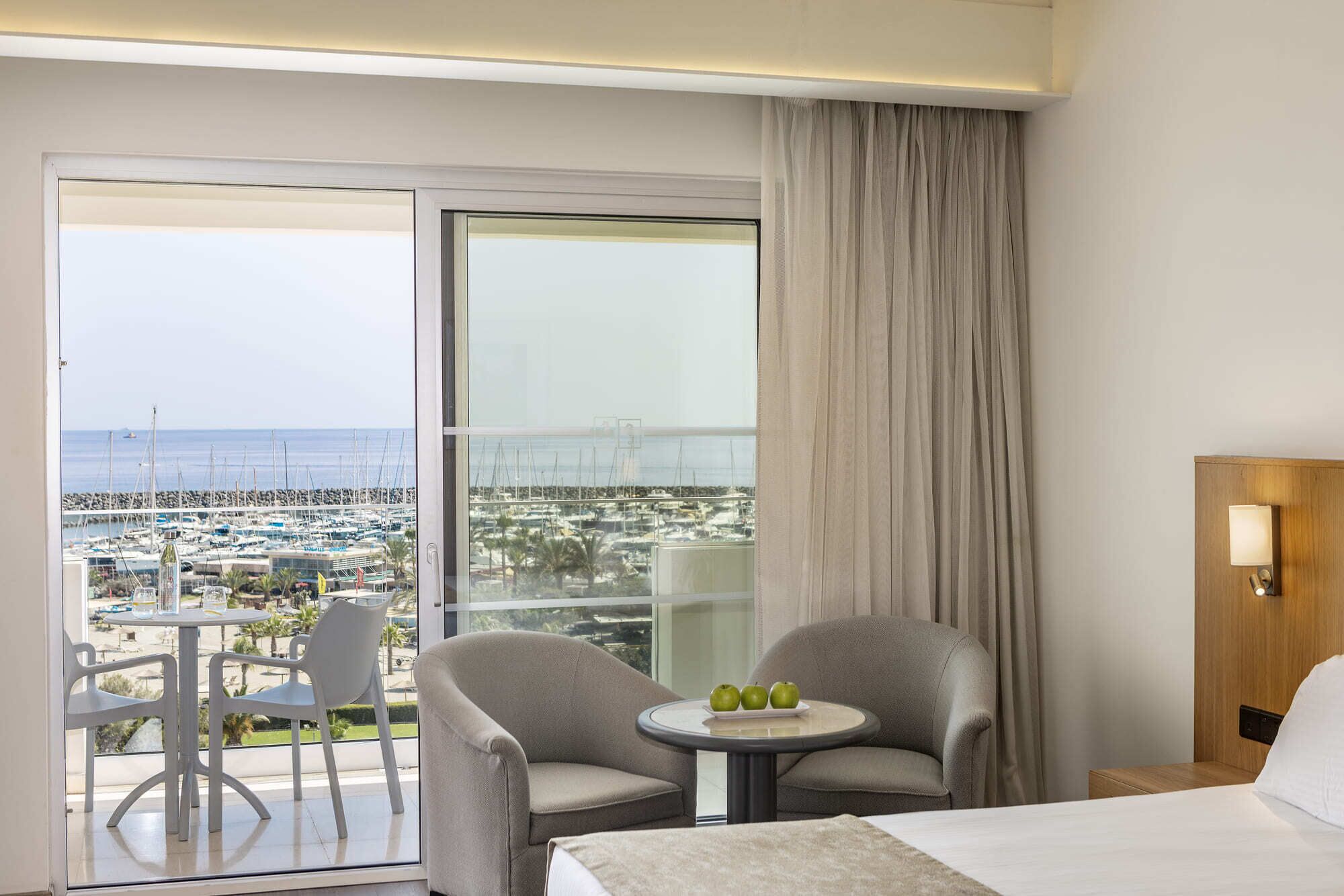 Habitación de hotel con balcón, dos sillas, una mesita con manzanas, cama y mesita de noche con lámpara.