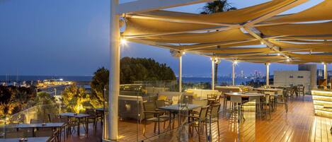 Un espacio exterior para comer con mesas y sillas, una terraza de madera y vistas del horizonte urbano al anochecer.