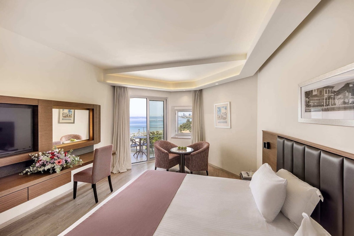Ein Hotelzimmer mit Bett, Fernseher, Schreibtisch, Stuhl, Tisch und einem Balkon mit Blick.