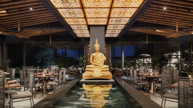 Un'area pranzo tranquilla con una statua centrale di Buddha, circondata da tavoli e sedie, sotto un soffitto decorato.