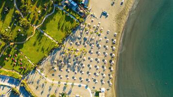 An aerial view of a beach resort with rows of umbrellas, a pool area, and a beachfront path.
