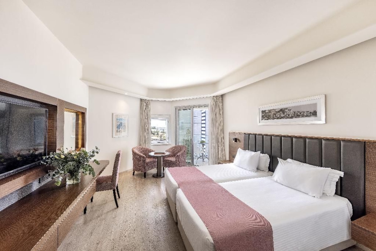 Ein Hotelzimmer mit Bett, Fernseher, einer Sitzecke mit Tisch und Stühlen sowie einem Fenster mit Vorhängen.